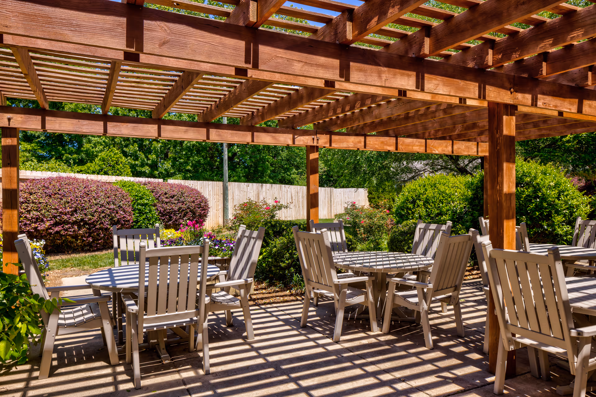 Outdoor patio area with wooden pergola providing partial shade over several round tables and chairs. The patio is surrounded by green bushes, colorful flowers, and a wooden fence in the background under a clear blue sky.