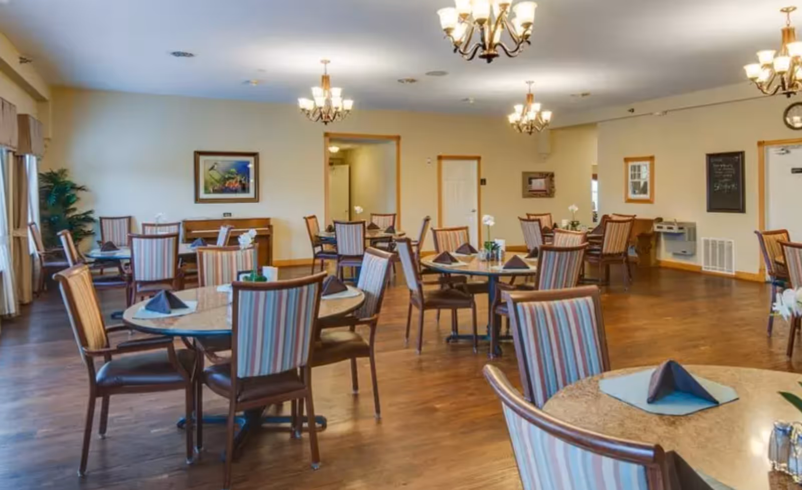 Spacious dining room with round tables and striped chairs set with folded napkins under chandeliers.