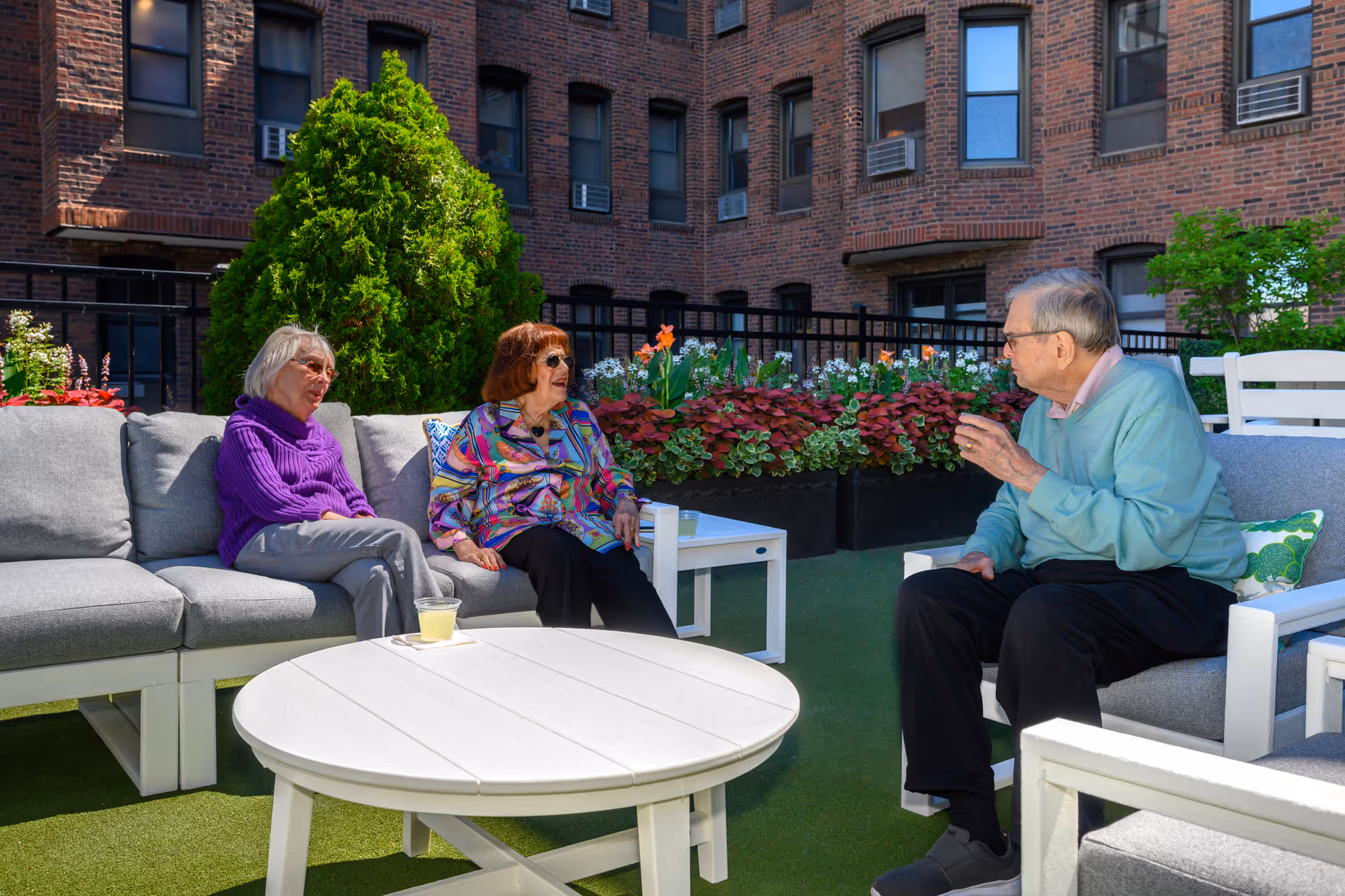 Three elderly people sitting and chatting on outdoor patio furniture in a courtyard with brick building walls and greenery in the background.