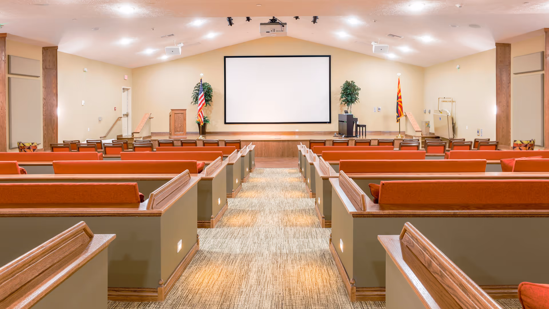 Interior view of a large meeting or presentation room with rows of wooden pew-style seating with red cushions facing a stage. The stage has a podium, two flags, two potted plants, a piano, and a large blank projection screen on the wall. The room is well-lit with ceiling lights and has a carpeted aisle down the center.