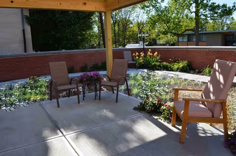 Outdoor patio area with a wooden pergola overhead, featuring three chairs and a small table with a flower pot. Surrounding the patio are flower beds with various plants and flowers, and a brick wall encloses the space. Trees and buildings are visible in the background.