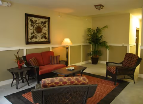 A cozy seating area with a red cushioned loveseat, two patterned armchairs, a coffee table, and a side table with a lamp and flower arrangement. There is a decorative wall hanging above the loveseat and a potted plant in the corner of the room.