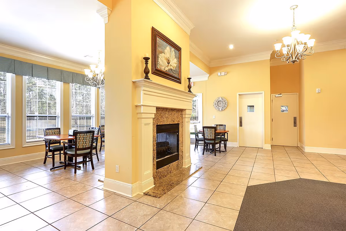 Interior view of a senior living facility dining area with large windows, several tables and chairs, a fireplace with a floral painting above it, and two chandeliers hanging from the ceiling. The walls are painted yellow and the floor is tiled.