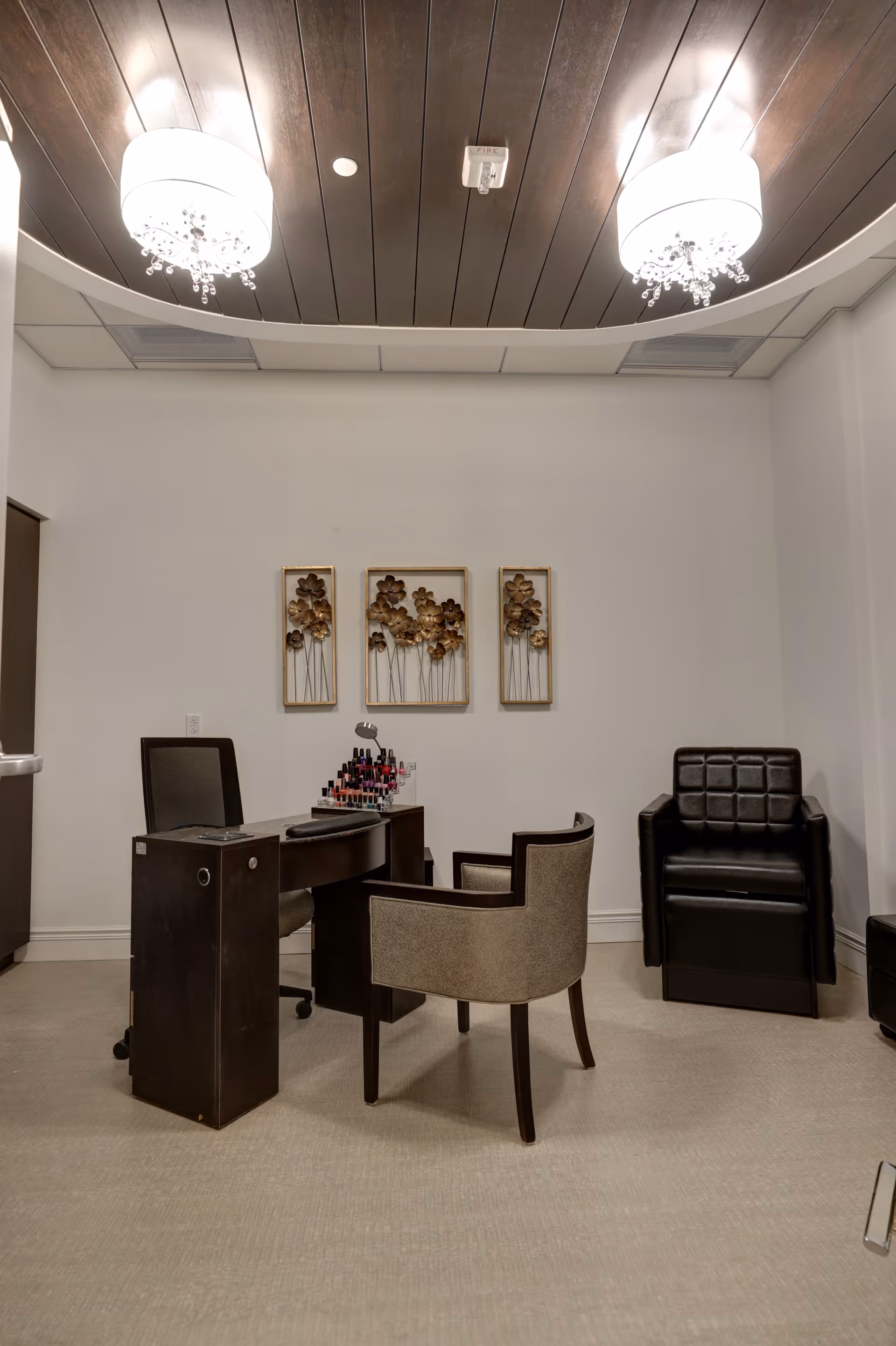 A small salon-style room with a manicure table, two chairs, a rack of nail polish, decorative wall art, and ceiling chandeliers.