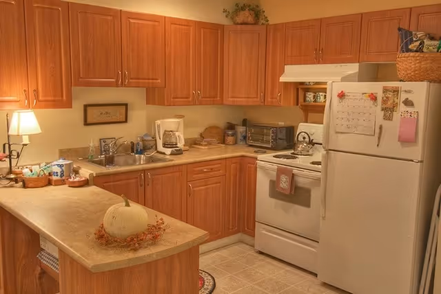 A cozy kitchen with wooden cabinets, a white refrigerator covered with magnets and notes, a white stove with a kettle on it, a toaster oven, a coffee maker, and a sink. There is a small lamp on the counter, a white pumpkin with autumn decorations on the island countertop, and various kitchen items neatly arranged.