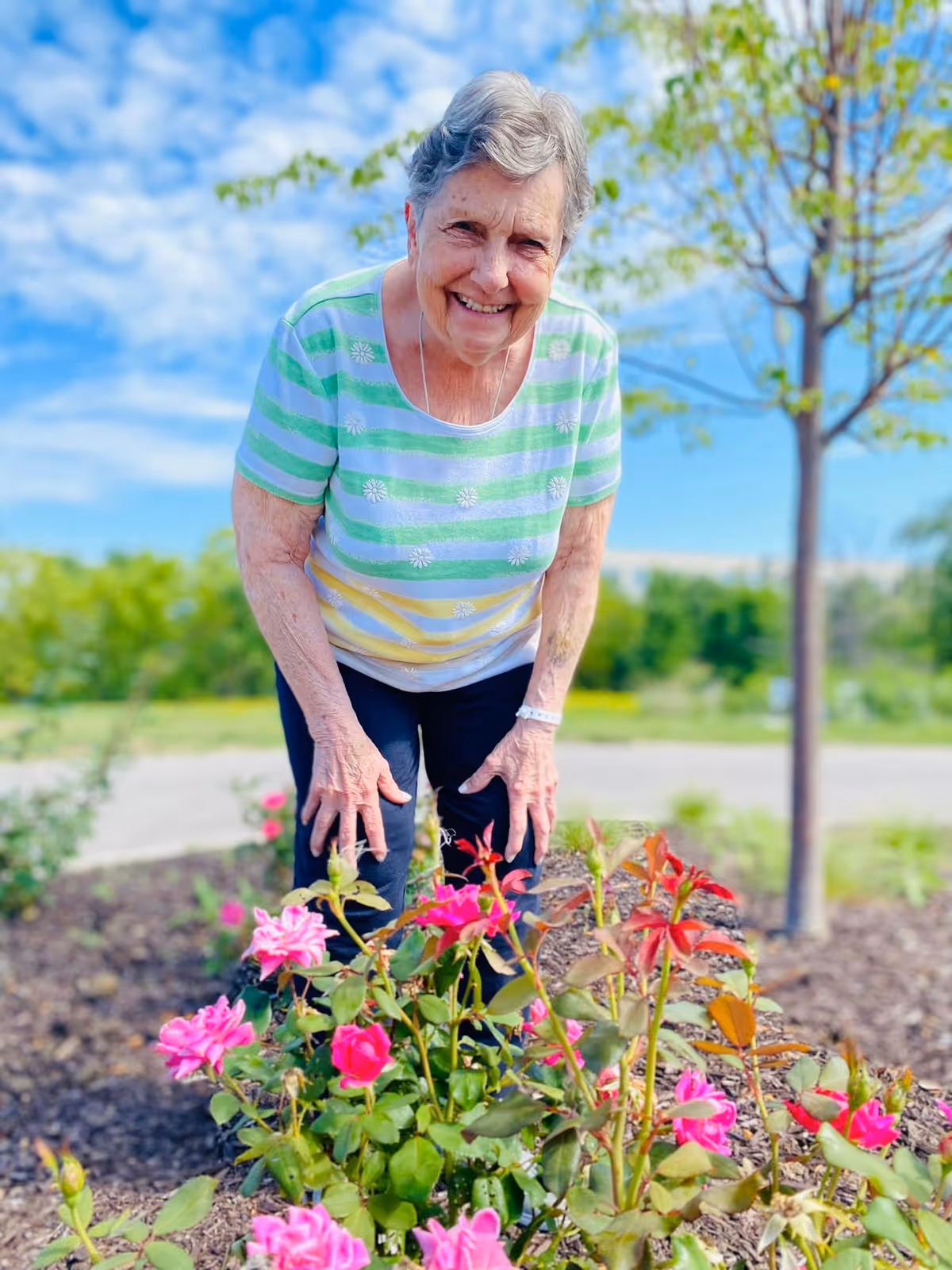 An elderly woman wearing a striped green, white, and yellow shirt and dark pants is smiling and bending over to look at pink flowers in a garden. The background shows a tree, greenery, and a partly cloudy blue sky.