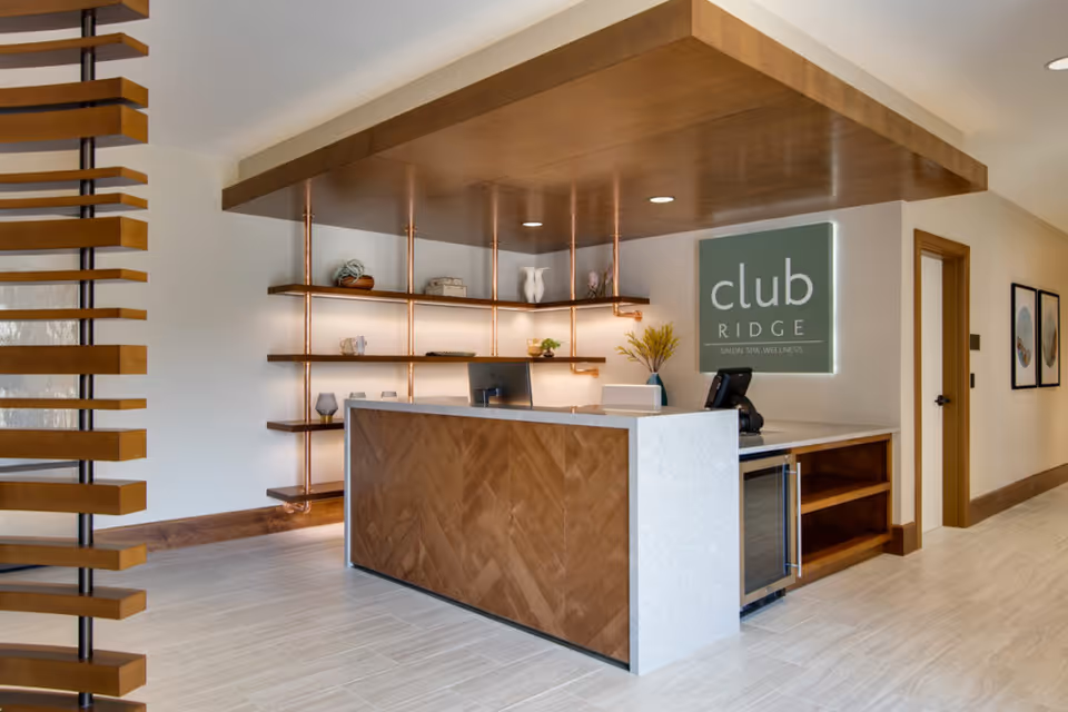 Reception desk area with modern wooden and white stone design, shelves with decorative items, and a sign on the wall that reads 'club RIDGE SALON SPA WELLNESS'. The space has light-colored flooring and walls with wooden trim, and a doorway and framed pictures are visible in the background.