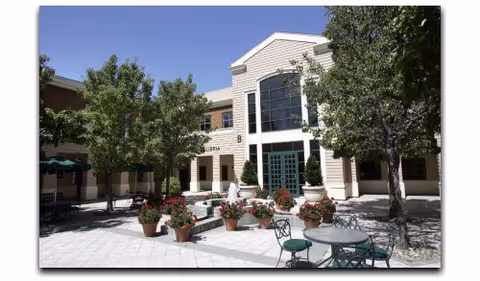 Sunlit courtyard with potted flowers, a small fountain and patio tables in front of the entrance to a senior living building.