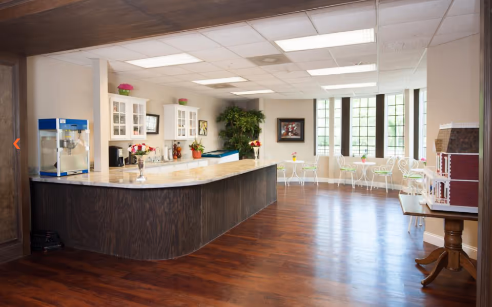 A bright and spacious room with a large marble countertop island, white cabinets on the wall, a popcorn machine, and several small white tables with chairs near large windows letting in natural light. There are decorative plants and flowers on the counter and tables, and a framed picture on the wall. A detailed dollhouse model sits on a wooden table to the right.