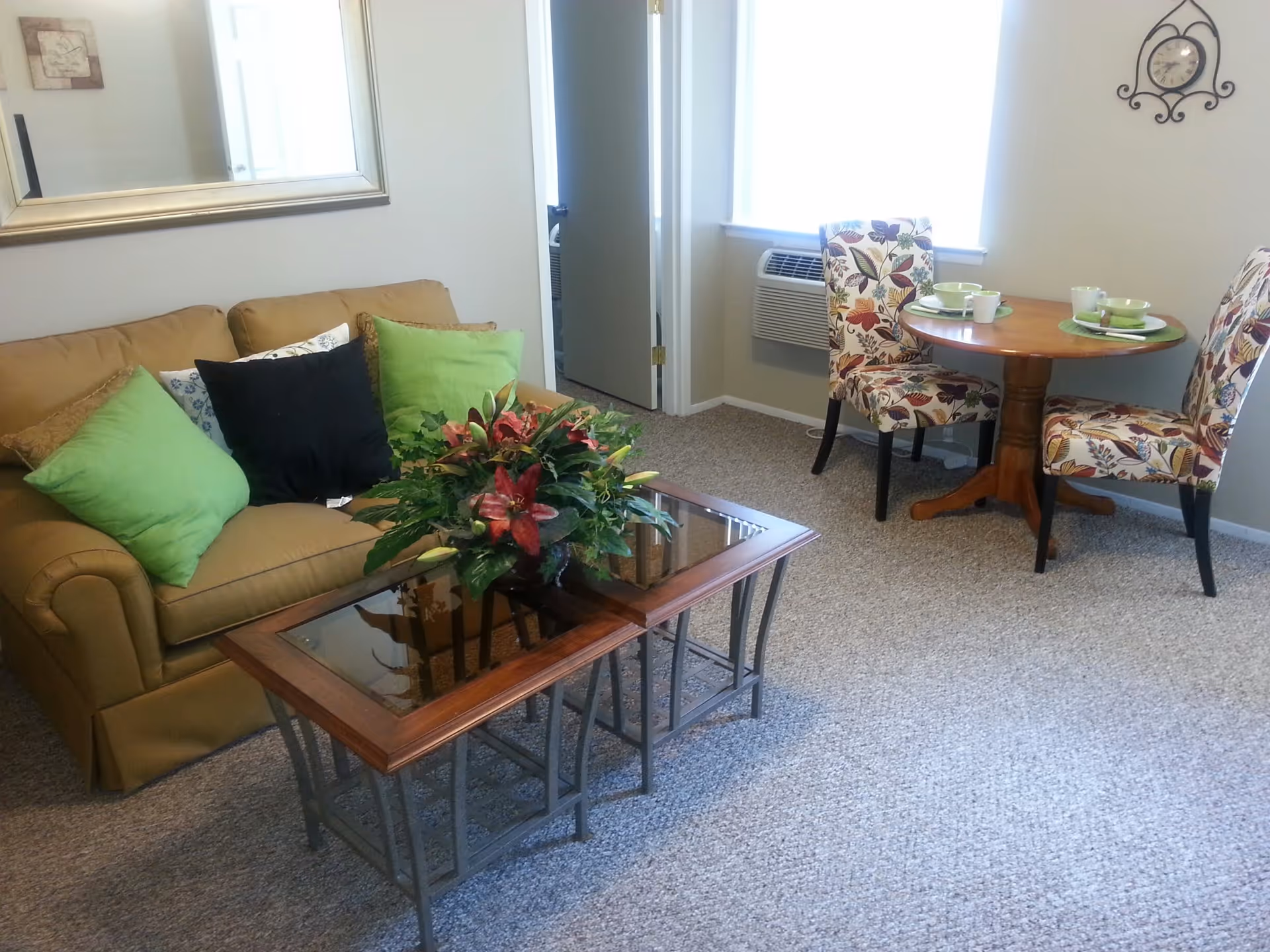 A cozy living room area with a tan sofa adorned with green, black, and patterned pillows. In front of the sofa is a glass-top coffee table with a floral arrangement. To the right, there is a small round wooden dining table set for two with floral upholstered chairs. A wall clock is mounted above the dining area, and a window with an air conditioning unit is visible behind the table.