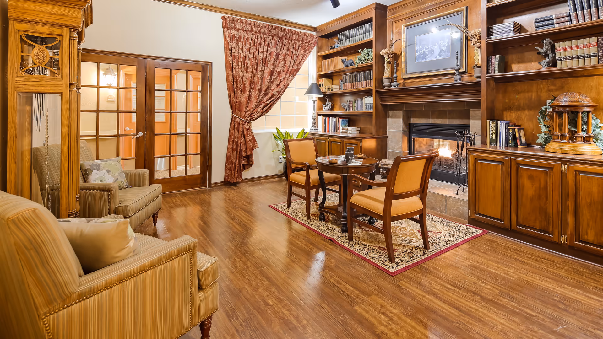 A cozy living room with wooden flooring, a fireplace surrounded by built-in wooden bookshelves filled with books and decorative items, a small round table with two chairs on a patterned rug, two armchairs with cushions, a large grandfather clock, and a window with red patterned curtains.