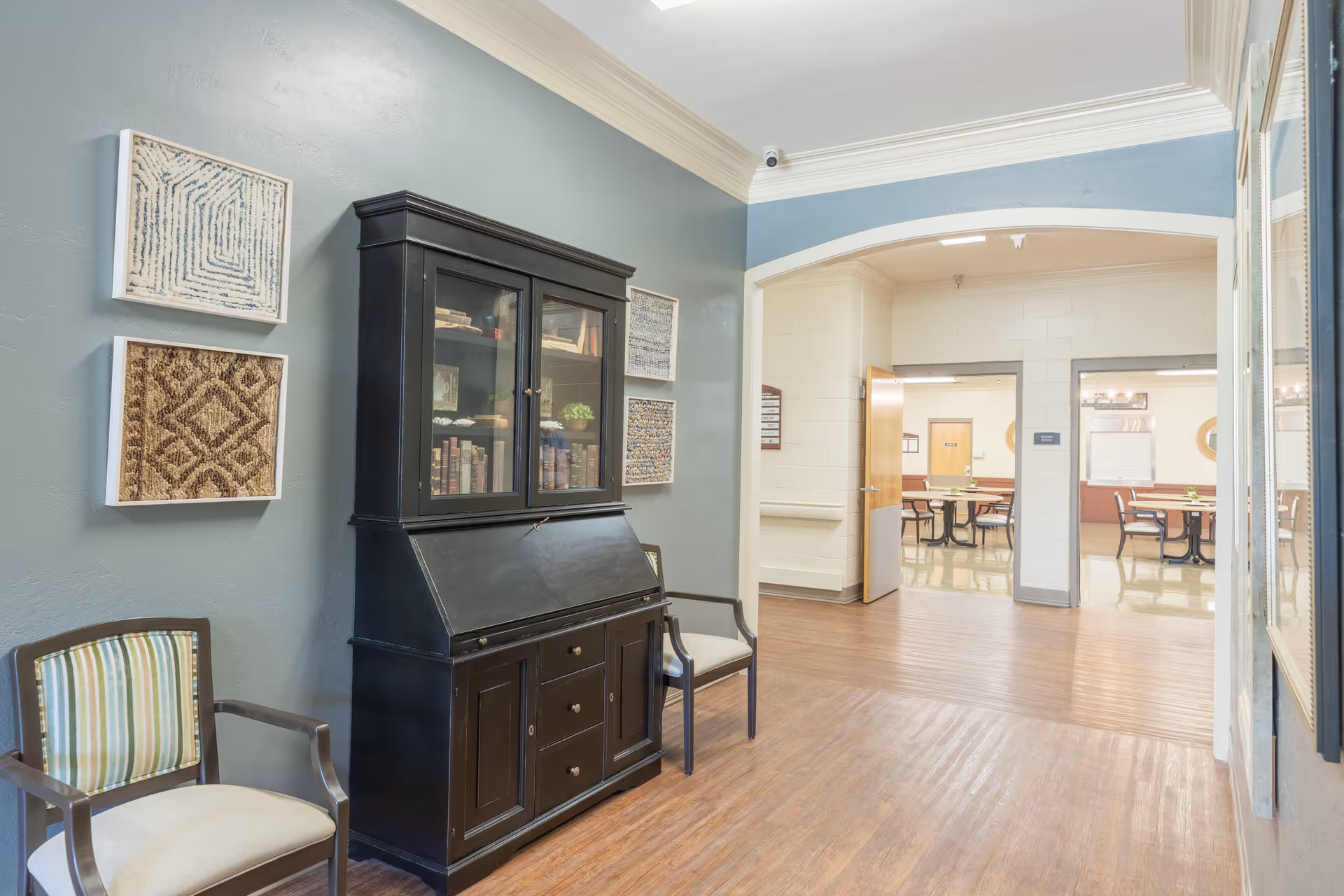 Interior hallway of a senior living facility with wooden flooring, a black cabinet with glass doors containing books, two chairs with striped cushions, and framed artwork on the wall. The hallway leads to a dining area with tables and chairs visible through an open archway.