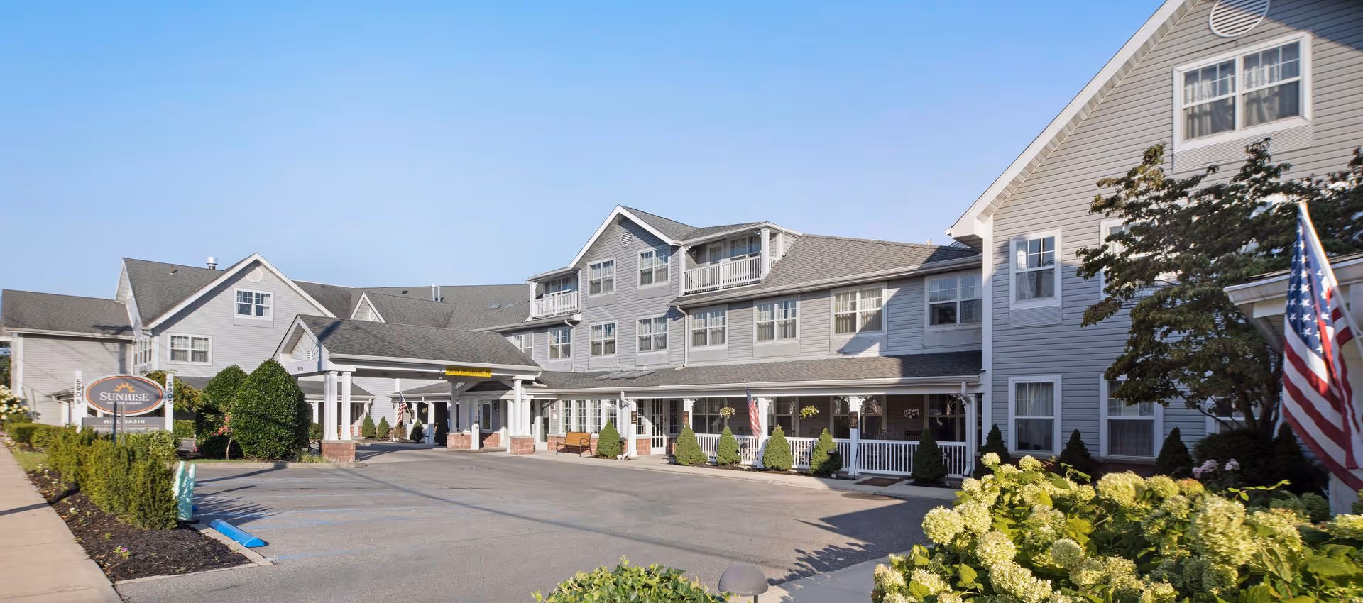 Front entrance of a gray multi-story senior living building with a covered drive, landscaped grounds, and an American flag.