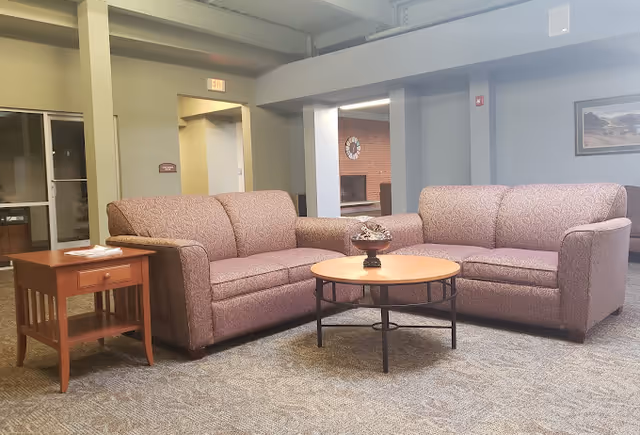 A lounge area with two matching upholstered sofas facing a round coffee table and a wooden side table in a carpeted common room.