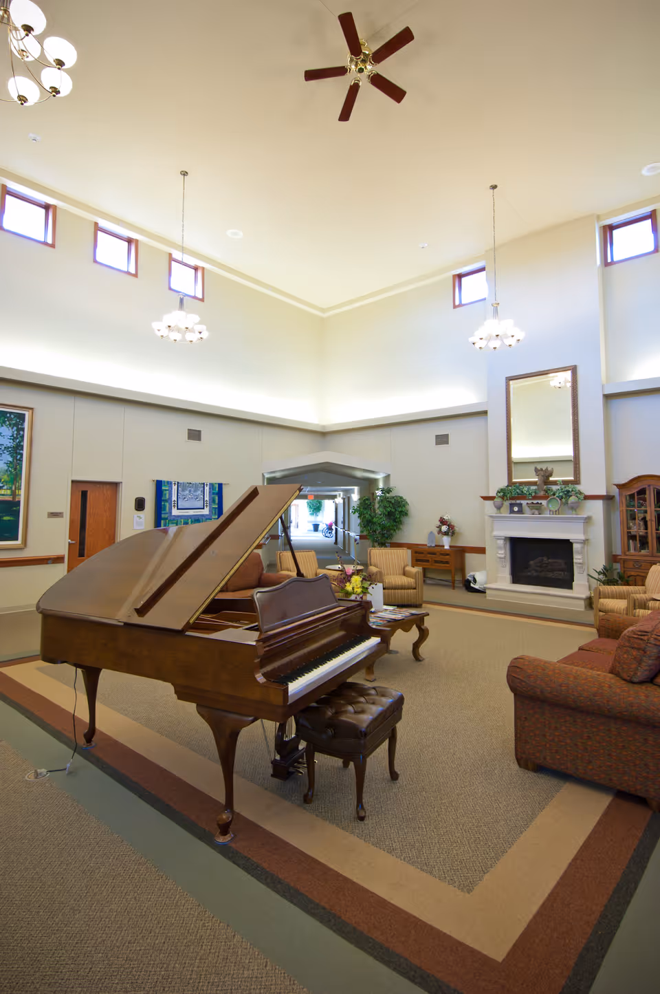 A spacious and well-lit living room area in a nursing and rehab center featuring a grand piano with a matching bench in the foreground. The room has high ceilings with ceiling fans and hanging light fixtures. There are several upholstered chairs and sofas arranged around a coffee table, and a fireplace with a large mirror above it. The walls have small windows near the ceiling and some framed artwork. A doorway leads to another area of the facility.