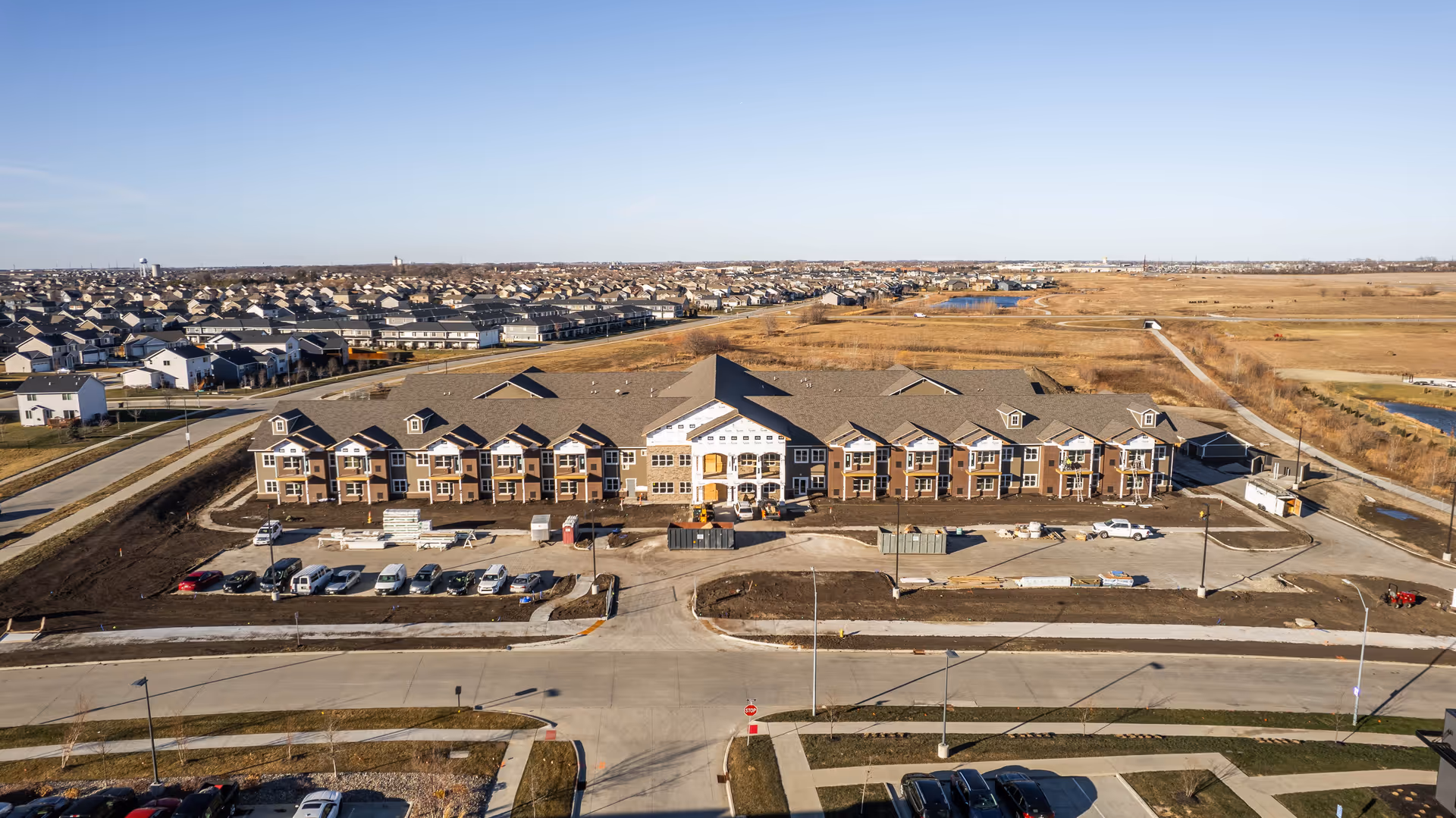 Aerial view of a two-story senior living building under construction with parked cars and surrounding suburban landscape.