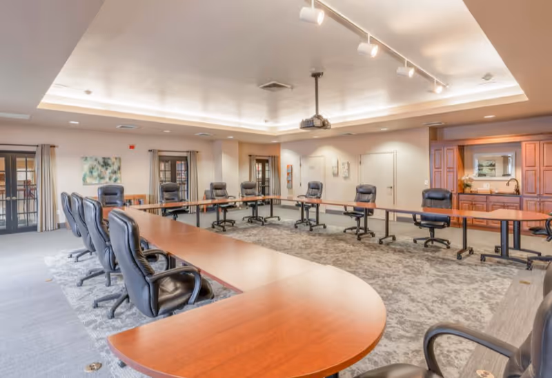 A large conference room with a U-shaped arrangement of wooden tables and black leather office chairs on wheels. The room has a carpeted floor, beige walls, a ceiling-mounted projector, and recessed lighting. There are double glass doors with curtains on one side and a wooden cabinet with a sink and mirror on the other side.