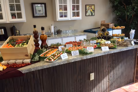 A buffet-style food spread on a long counter with various appetizers including vegetables, cheese, crackers, and small skewers. The counter is in a room with white cabinets and framed pictures on the wall. There are also bottles of oil or vinegar and a tiered tray with more food items.