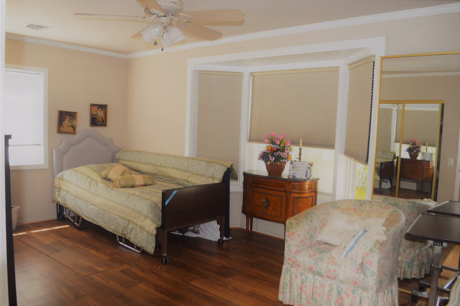 A cozy bedroom with a single bed covered in a patterned bedspread, a floral upholstered armchair with a pillow, a wooden side table with a flower pot and framed photo, and a large mirror reflecting part of the room. The room has wooden flooring, beige walls, and windows with beige blinds.