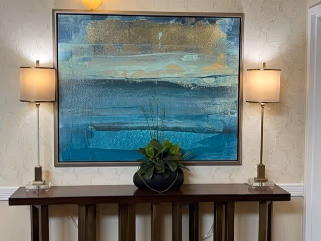 A wooden console table with a black vase holding green plants placed in the center. Above the table hangs a large abstract painting with shades of blue, teal, and gold. On either side of the painting, there are two wall-mounted lamps with beige lampshades. The wall behind has a subtle textured pattern.