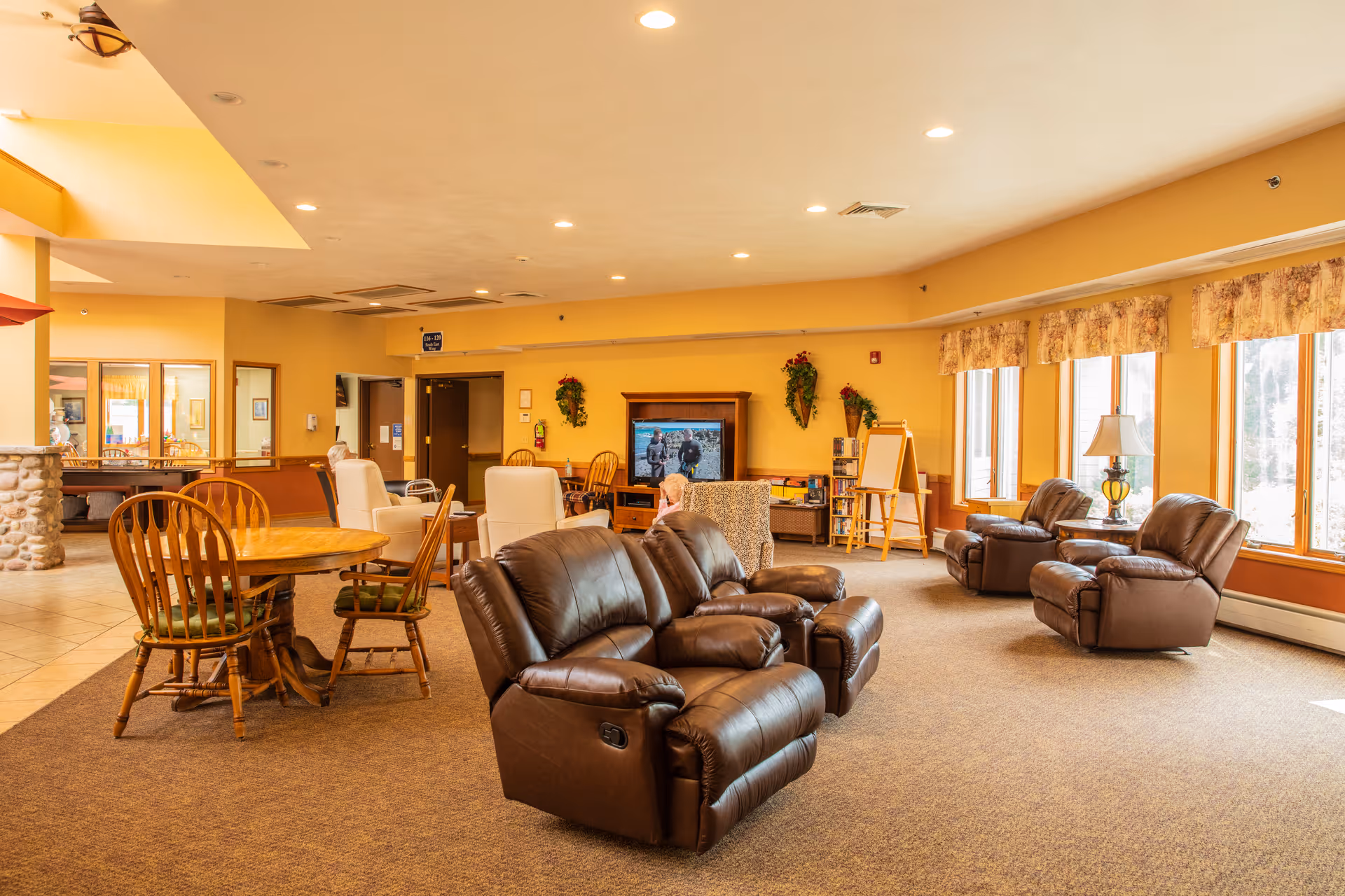 A spacious living room area in Oakwood Manor with several brown leather recliners, wooden chairs around a round table, large windows with floral curtains letting in natural light, and a television mounted on a wooden stand. The walls are painted yellow and decorated with greenery and flowers.
