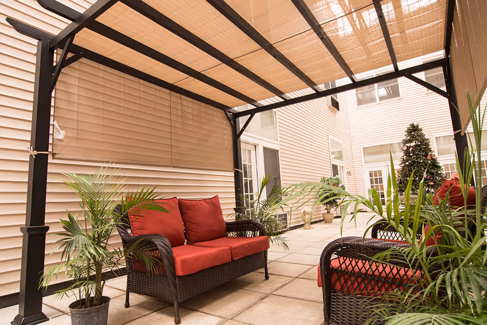 Covered outdoor patio area with wicker furniture featuring red cushions, potted plants, and a Christmas tree in the background, adjacent to a beige building with multiple windows and doors.