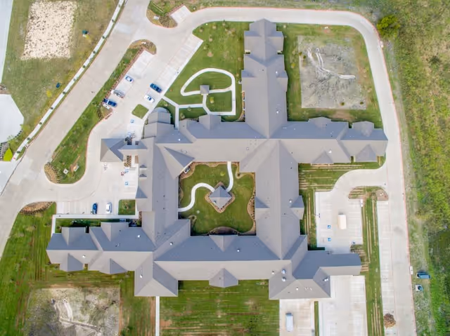 Aerial view of Legend Oaks Healthcare and Rehabilitation - Waxahachie showing the building's roof layout, surrounding parking lots, driveways, and landscaped green areas with pathways.