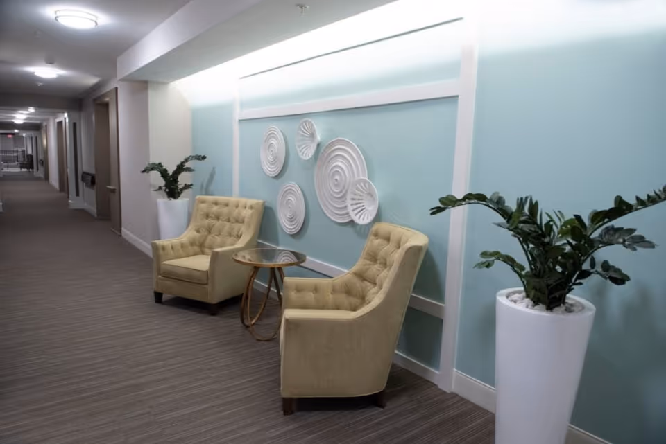 A hallway seating area in a senior living facility with two beige tufted armchairs and a small round glass-top table between them. The wall behind the chairs is painted light blue and decorated with white circular wall art. There are two tall white planters with green plants on either side of the seating area. The hallway extends into the distance with doors and ceiling lights visible.
