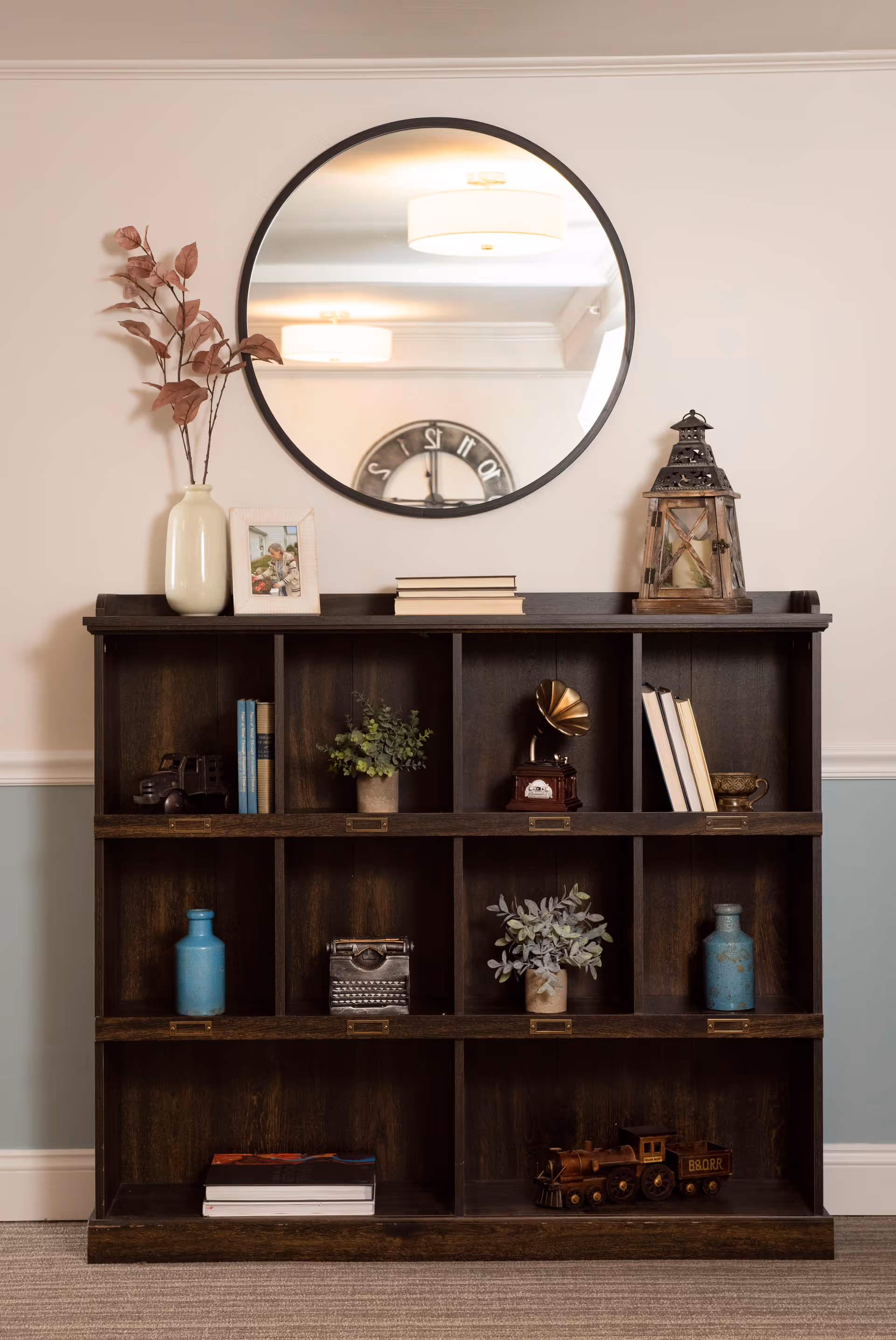 A wooden cubby bookshelf with various decorative items including books, plants, a vintage typewriter, a model train, a gramophone, a lantern, and vases. Above the bookshelf is a round mirror reflecting ceiling lights and a wall clock.