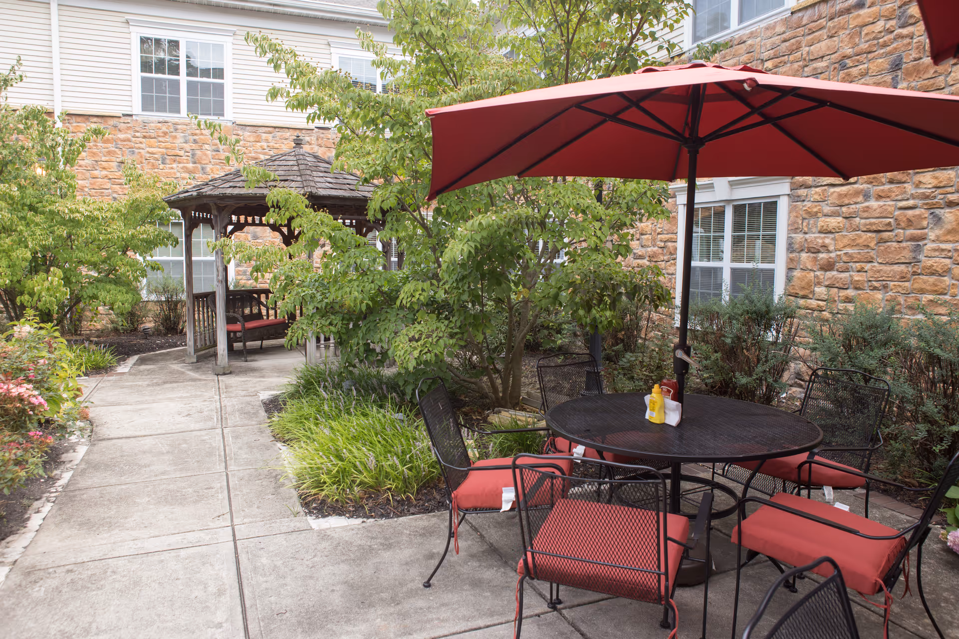 Outdoor patio area with a round black metal table and six chairs with red cushions. A large red umbrella shades the table. Surrounding the patio are green bushes and trees, with a stone building wall and windows in the background. A wooden gazebo with a bench is visible along a concrete pathway.