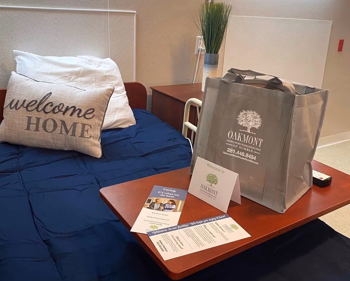 A neatly made bed with a navy blue blanket and a pillow that says 'welcome HOME'. Next to the bed is a wooden table with a gray tote bag featuring the Oakmont Healthcare & Rehabilitation Center of Humble logo and contact information. On the table are also brochures and a welcome card from Oakmont. A small plant is placed on a nightstand behind the table.