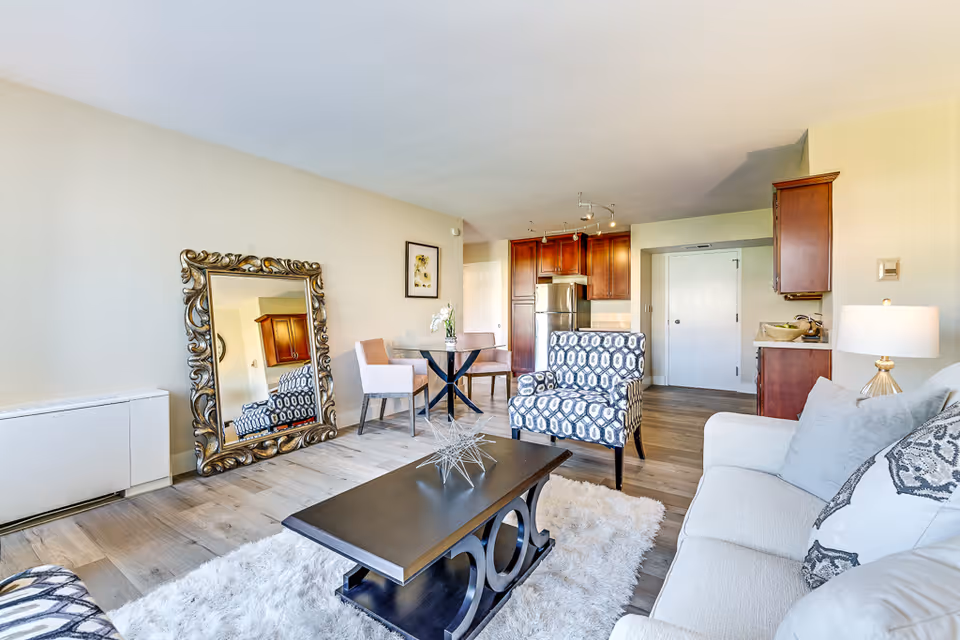 A bright and modern living room area in a senior living facility featuring a white sofa with patterned pillows, a patterned armchair, a dark wooden coffee table on a white shag rug, a large ornate floor mirror leaning against the wall, and a small dining table with two chairs near a kitchen with wooden cabinets and stainless steel refrigerator.