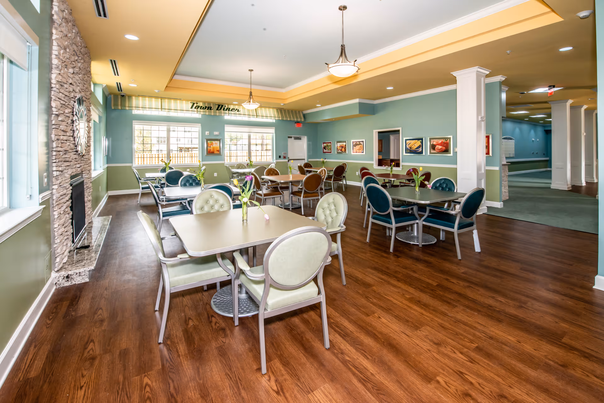 A bright and spacious dining room with several tables and chairs arranged neatly. The room features wood flooring, a stone fireplace on the left wall, and large windows letting in natural light. The walls are painted in a soft green with white trim, and there are framed pictures on the walls. A sign above the windows reads 'Town Diner'.