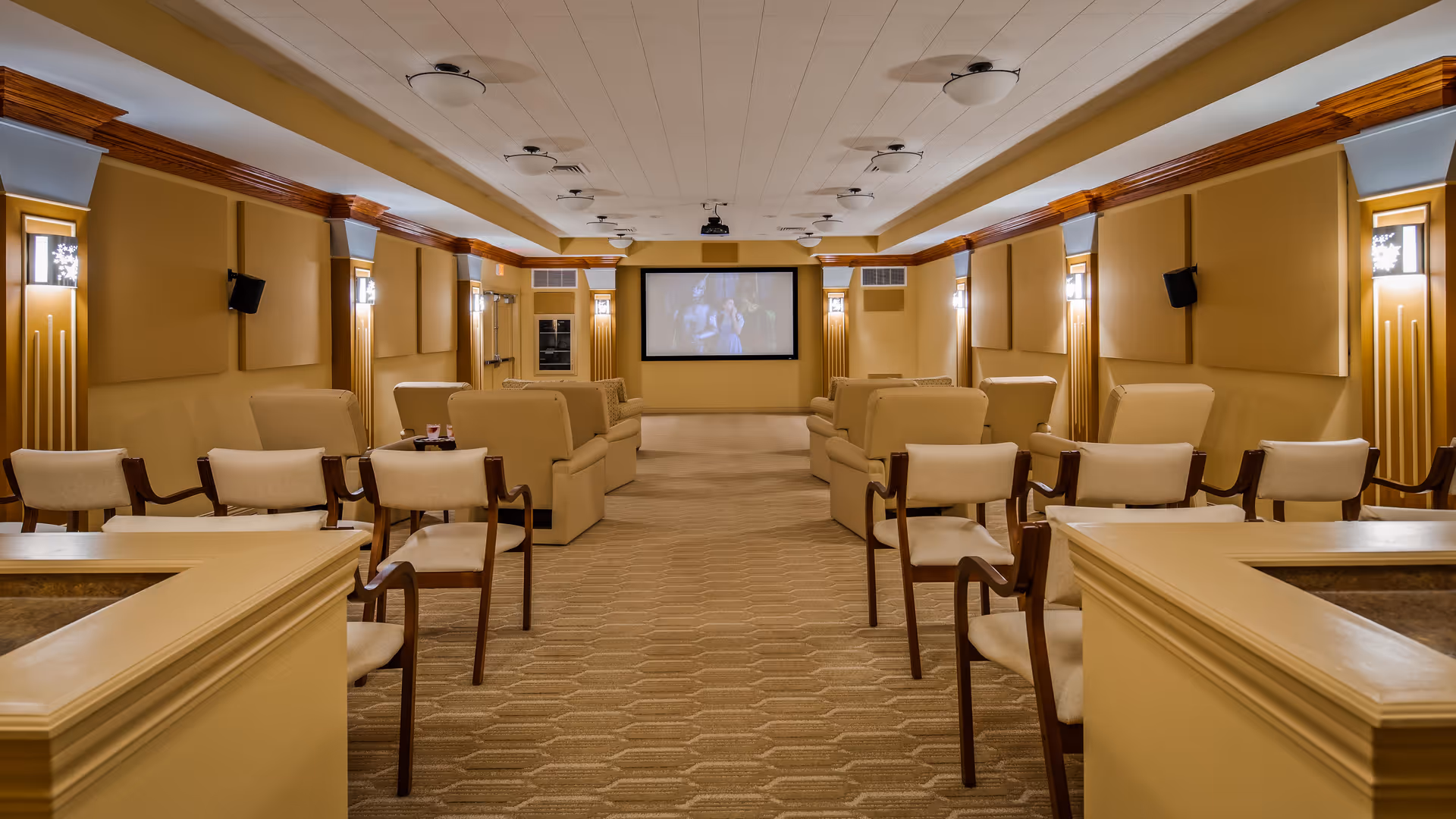 A cozy theater room with beige walls and carpet, featuring multiple rows of cushioned chairs and sofas facing a large screen at the front. The room is softly lit with wall sconces and ceiling lights, creating a warm and inviting atmosphere.