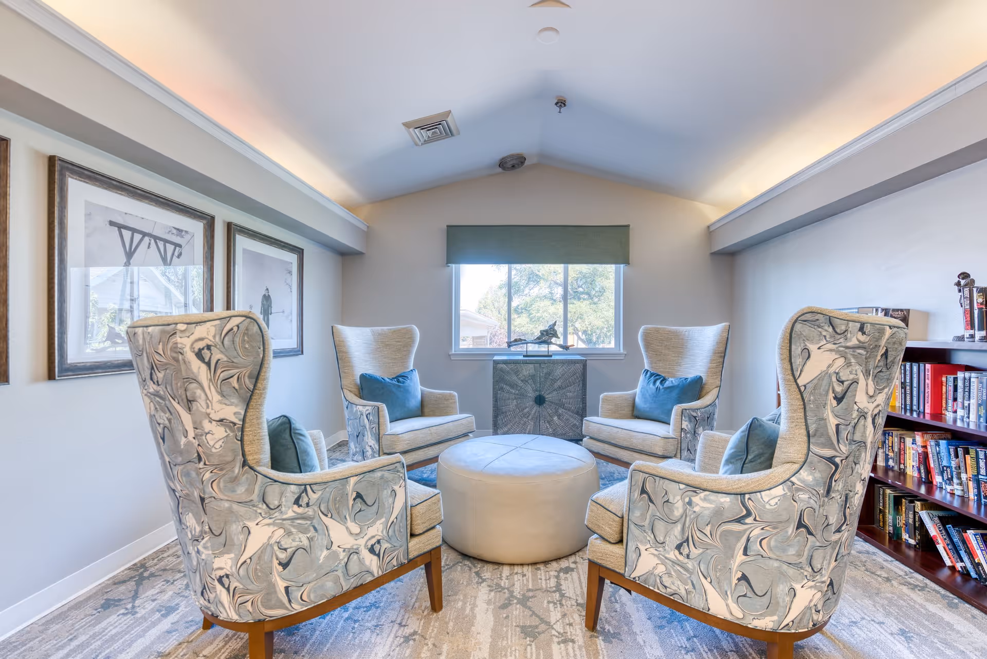 A cozy sitting area in a senior living facility with four patterned armchairs arranged around a round ottoman. The room has a window with a green valance, framed artwork on the wall, and a bookshelf filled with books. The ceiling is vaulted with recessed lighting.