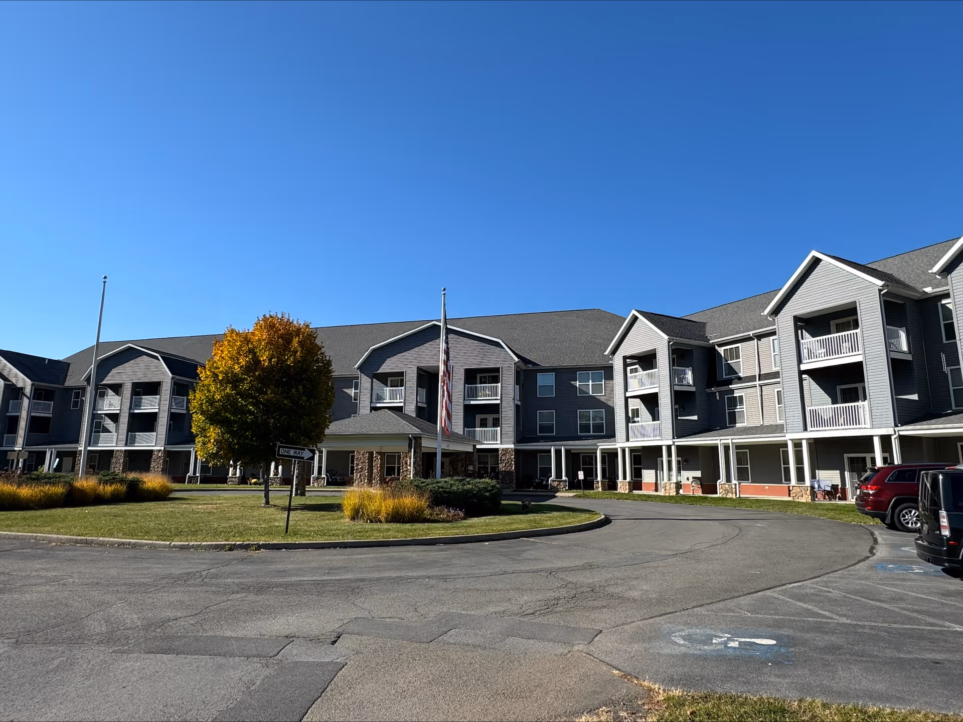 Exterior view of a multi-story senior living facility building with gray siding and white balconies under a clear blue sky. There is a circular driveway with a landscaped island featuring a tree with autumn-colored leaves and an American flag on a flagpole. Several parked cars are visible on the right side.