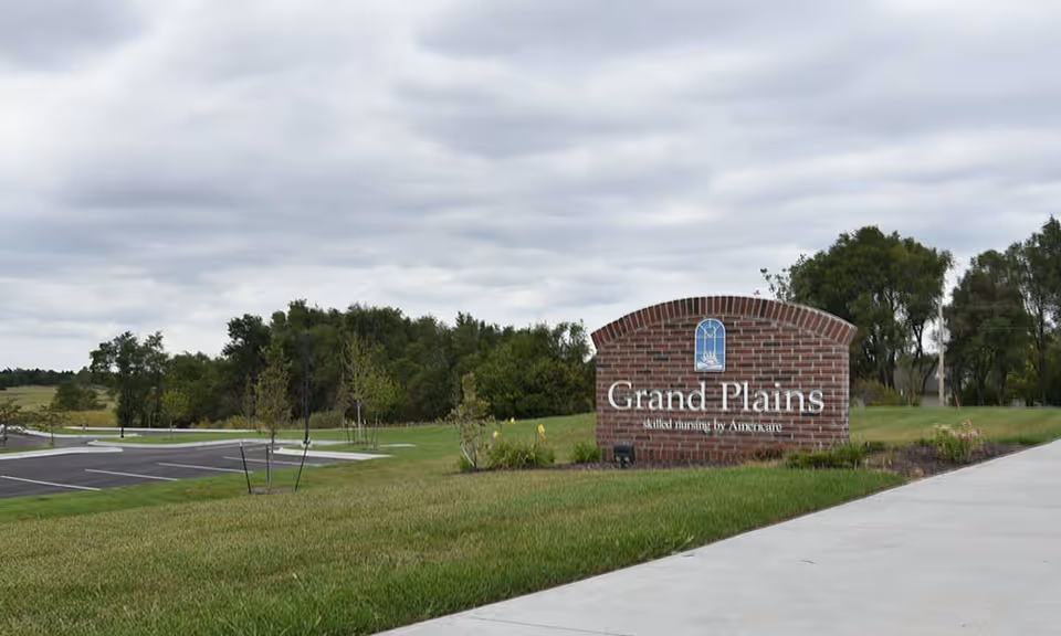 Outdoor view of the Grand Plains Skilled Nursing and Rehabilitation facility sign made of brick with a landscaped grassy area and trees in the background under a cloudy sky.