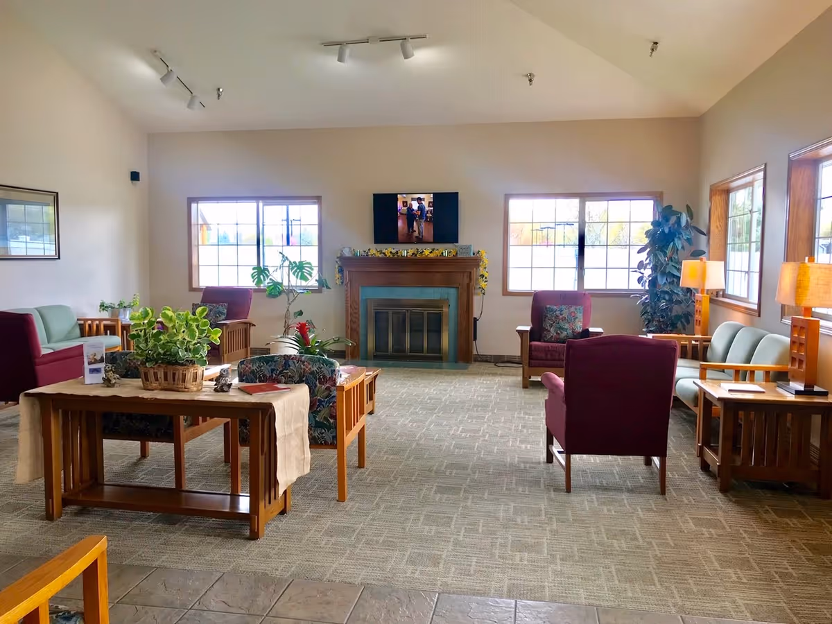 A bright and spacious living room area in an assisted living facility with multiple armchairs and sofas arranged around a central wooden table with a plant on it. There is a fireplace with a TV mounted above it, two large windows letting in natural light, and several lamps providing additional lighting. The room has a carpeted floor and light-colored walls.
