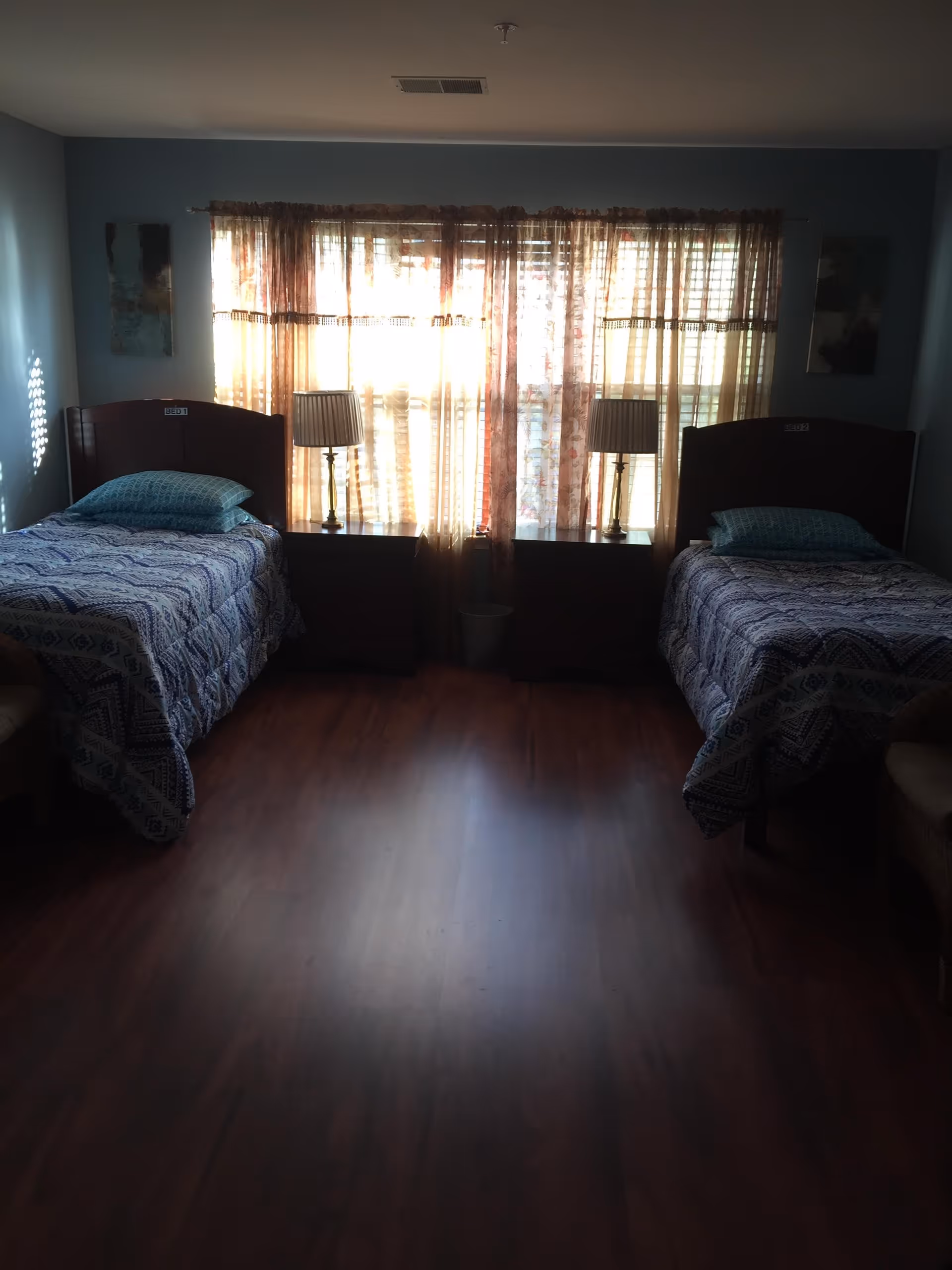 A dimly lit bedroom with two single beds placed on opposite sides of the room. Each bed has a patterned bedspread and a pillow. Between the beds are two nightstands, each with a lamp. A window with sheer curtains allows some natural light to filter into the room. The floor is wooden, and there are two chairs partially visible on either side of the beds.