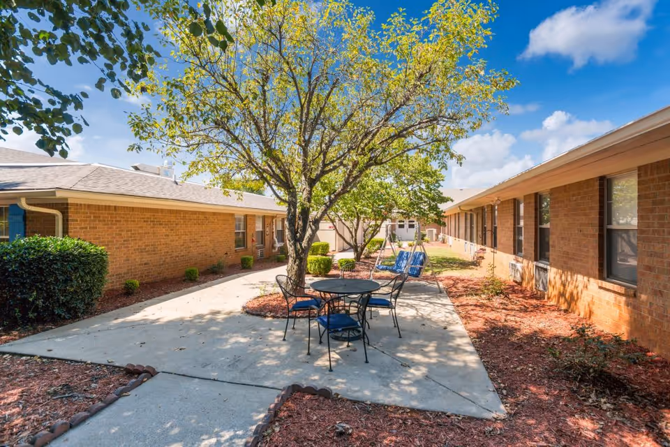 Outdoor courtyard area at Charter Senior Living of Bowling Green featuring a concrete patio with a round metal table and four chairs with blue cushions, surrounded by two single trees and landscaped with mulch and bushes. The courtyard is flanked by brick buildings under a blue sky with some clouds.