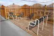 Outdoor patio area with wooden decking and a wooden privacy fence. The space includes several chairs and a small round table arranged for seating and relaxation.
