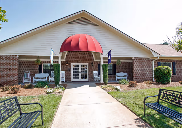 Front exterior view of a single-story brick building with a peaked roof and a red awning over the entrance. The entrance has double glass doors flanked by white columns and outdoor seating including white rocking chairs and benches. There are two flags on either side of the entrance and landscaped greenery along the walkway leading to the door.