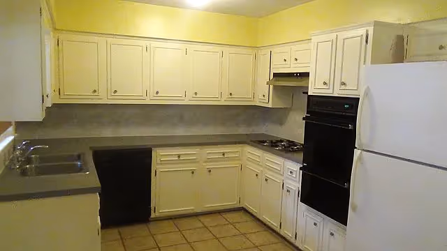 A kitchen with white cabinets and drawers, a double sink, a black dishwasher, a black built-in oven, a stovetop with a range hood, and a white refrigerator. The countertops are gray, and the floor is tiled with beige tiles. The walls are painted yellow above the cabinets.