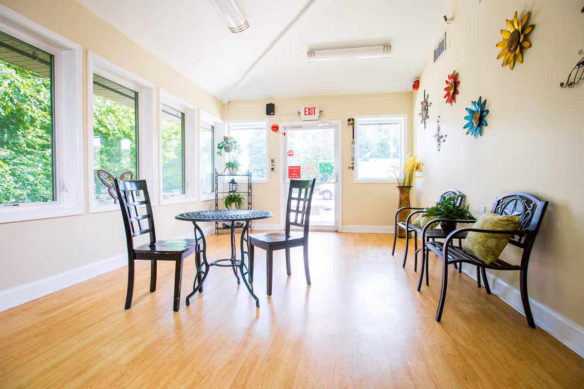 A bright room with large windows on the left side showing green trees outside. The room has light-colored walls and wooden flooring. There is a small round metal table with two black chairs in the center. On the right side, there are three black metal chairs with green cushions and a small table with a plant. The wall on the right is decorated with colorful metal flower wall art. At the far end, there is a glass door with an exit sign above it and two windows on either side.