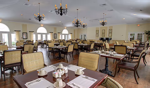 A spacious dining room with multiple tables and chairs arranged neatly. Each table is set with white cups, saucers, napkins, and condiments. The room features large arched windows allowing natural light, chandeliers hanging from the ceiling, and framed artwork on the walls. The floor is wooden, and the overall ambiance is warm and inviting.