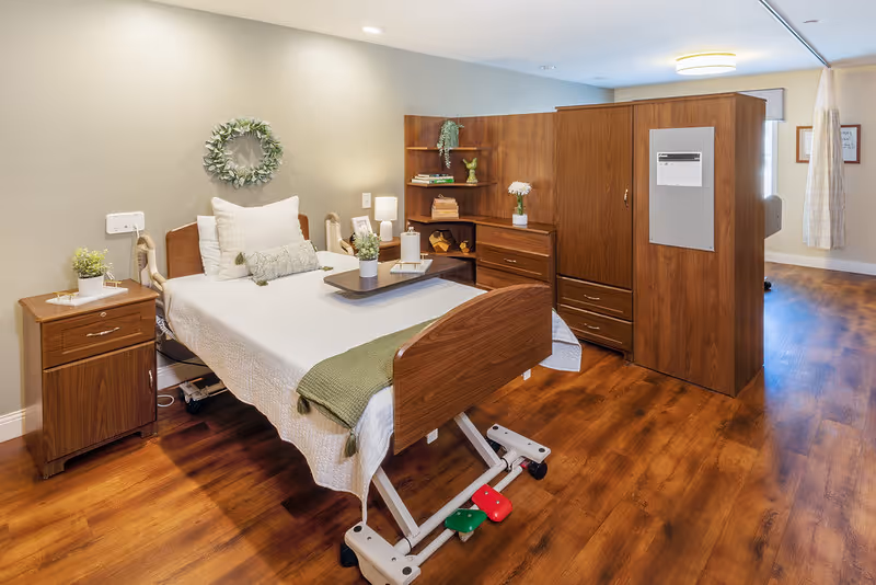 A well-lit senior living bedroom featuring a hospital-style adjustable bed with white bedding and green throw, a wooden nightstand with a small plant and lamp, and a large wooden wardrobe with shelves and drawers. The room has wooden flooring and neutral-colored walls with a decorative wreath hanging above the bed.