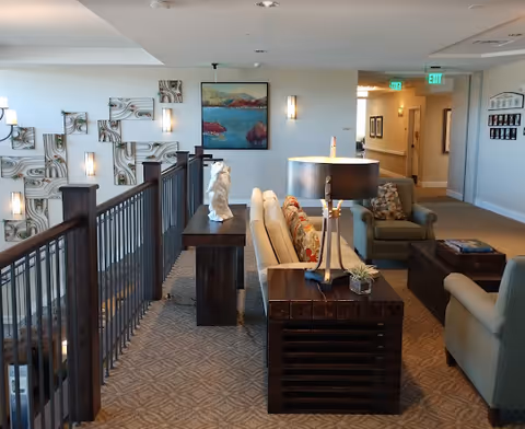 Interior view of a senior living facility lounge area with beige and green upholstered chairs, a wooden side table with a lamp and a small plant, decorative wall art, and a railing overlooking a lower level.