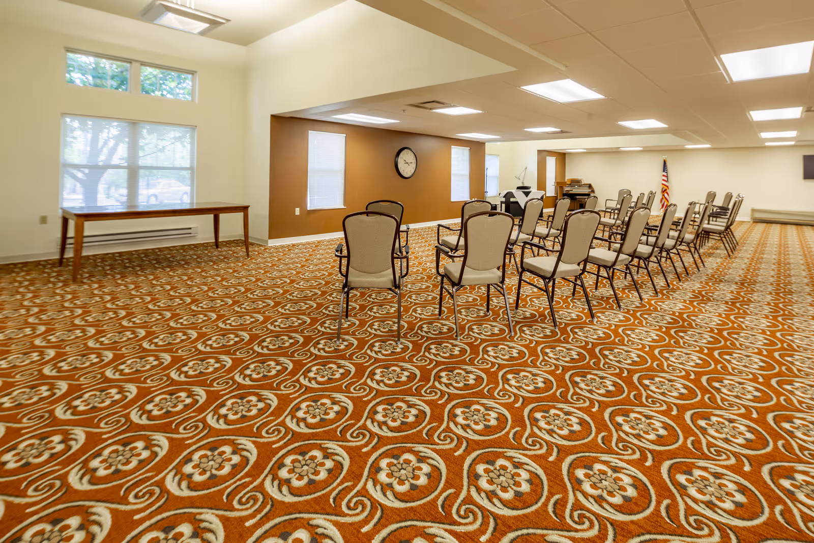 A large carpeted meeting room with rows of chairs, a piano, a wall clock and an American flag.