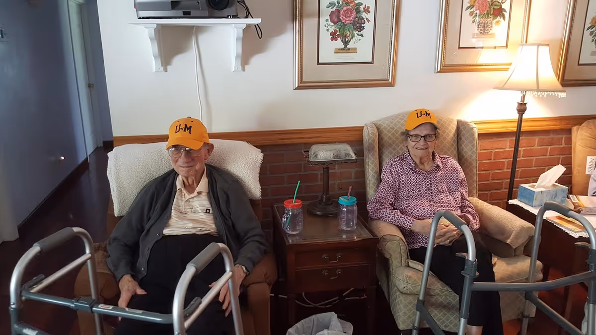 Two elderly individuals sitting in armchairs in a living room setting, each wearing a yellow cap with the letters 'U-M'. Both have walkers in front of them. There is a small wooden table between them with two cups and a lamp on it. The wall behind them has framed floral artwork and a floor lamp to the right.