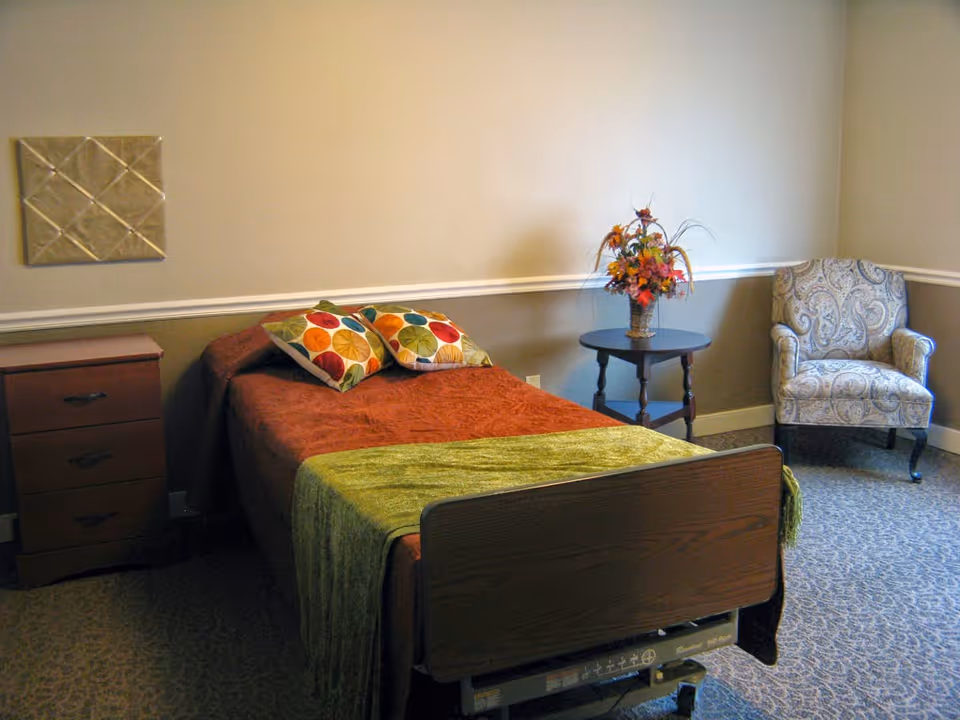 A bedroom in a senior living facility featuring a single bed with a brown bedspread and green throw, two colorful pillows, a wooden nightstand with three drawers, a round side table with a floral arrangement, and a patterned armchair in the corner. The walls are painted beige with a white chair rail molding.